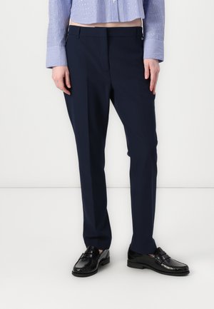 Pantaloni navy su misura con finitura liscia, caratterizzati da un taglio classico e una sottile piega frontale, indossati con scarpe in pelle nera.