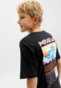 Blond jongetje lacht, kijkt naar links, draagt een zwart T-shirt met een kleurrijk Japans kasteel en teksten op de achterkant, tegen een effen witte achtergrond.