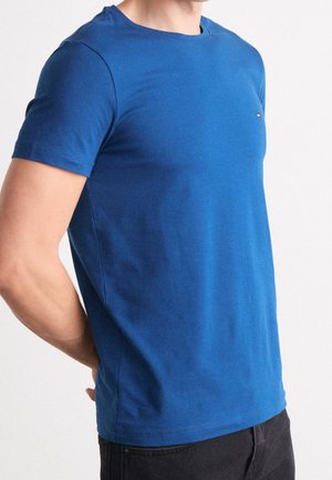 Homme portant un t-shirt bleu uni à manches courtes avec un petit logo sur la poitrine, associé à un pantalon noir, sur fond blanc.