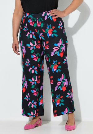 Plus size person iført sorte bukser med brede ben med farverigt blomstret mønster og lyserøde flade sko, står mod en lys væg.