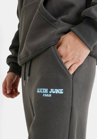 Grijze sweatshirt en broek met een losse pasvorm, zachte textuur en een front pocket, met blauwe tekst "SIXTH JUNE PARIS" op het been.