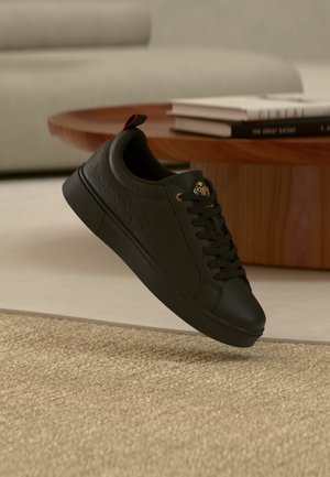 Schwarzer Sneakers mit niedrigem Schnitt, geprägtem Muster, goldenen Ösen und farbigem Logo auf der Zunge, nahe einem Holztisch und beigem Teppich platziert.