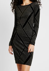 Svart stickad klänning med långa ärmar, rund halsringning och ett geometriskt mönster av glänsande silver och svarta diagonala ränder.