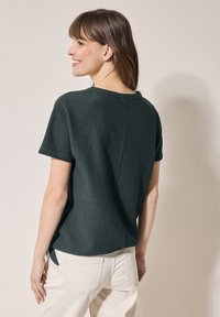 Chemise à manches courtes texturée vert foncé avec un col rond et des fentes sur les côtés, associée à un pantalon de couleur claire.