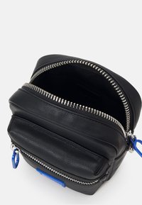 Crna kožna torba s dvostrukim zatvaračem, s teksturiranom površinom, srebrnim dodacima i plavim akcentom na prednjem džepu.