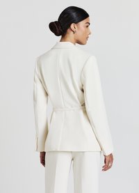 Blazer bianco su misura con silhouette strutturata, maniche lunghe, dettaglio con fiocco sul retro e texture liscia. Indossato sopra pantaloni abbinati.