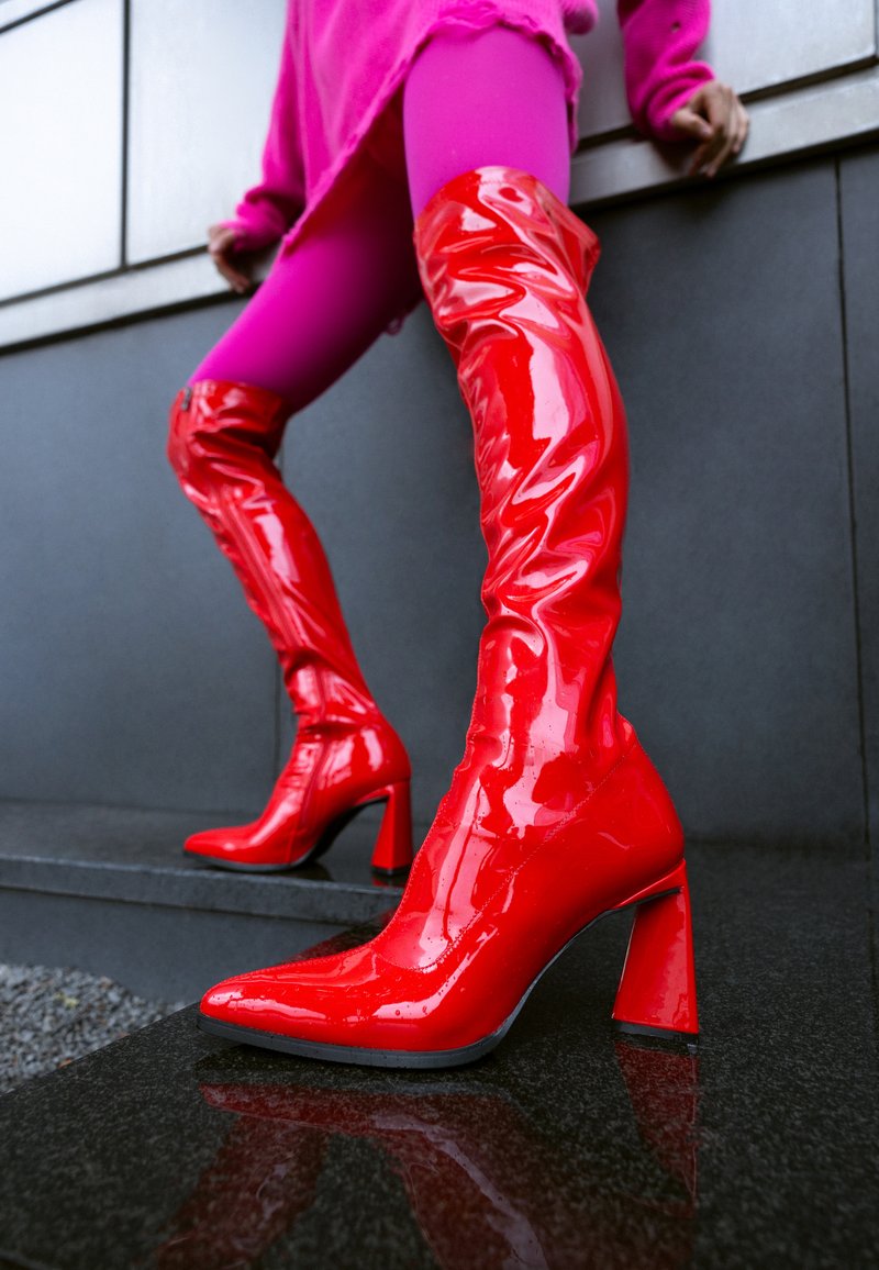 Botas de cuero sintético rojo brillante hasta el muslo con punta afilada, acabado brillante y tacón grueso alto. Superficie texturizada con ligeras arrugas.