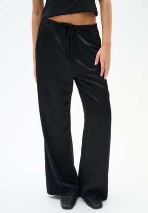 Zwarte wijde broek met een gladde, glanzende textuur, met een elastische tailleband en koord. Draagt zwarte schoenen.
