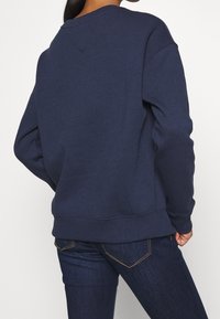 Persona con felpa blu scuro e jeans blu scuro, vista da dietro con una mano appoggiata sull'anca.