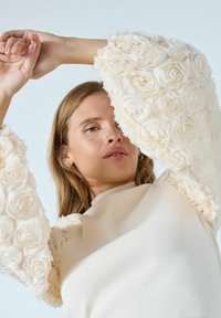 Kreemjas topp, millel on õhuliste varrukate peal lillekujulised tekstuuriga kaunistused. Sile kangas, mis alumises osas vormib keha ja millel on soonikuga krae. Pehme ja kerge välimus.