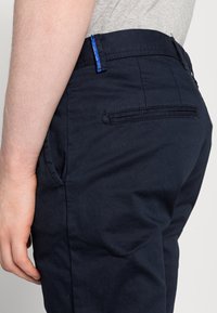 Marineblaue Baumwollhose mit glatter Textur, ausgestattet mit einer Gesäßtasche und einem blauen Etikett am Bund, aus seitlicher Perspektive gezeigt.