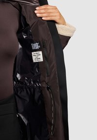 Giacca imbottita con esterno nero e fodera marrone, caratterizzata da un colletto in pelliccia sintetica, etichette interne e un cordoncino regolabile all'orlo.