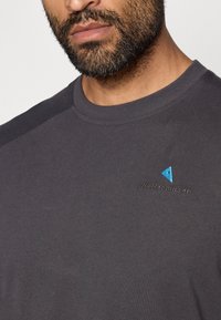 T-shirt de manga curta cinza-escuro com decote redondo, apresentando um logótipo triangular azul bordado e o nome da marca no peito. Textura suave.