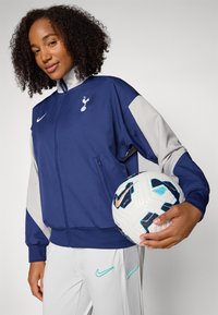 Blå och grå idrottsjacka med dragkedja och logotyp, i kombination med ljusgrå byxor, håller en mönstrad fotboll, mot en vit bakgrund.