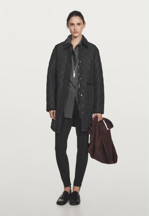 Sort quiltet frakke med en struktureret krave, båret over en langærmet grå skjorte. Sorte slim-fit bukser og skinnende loafers. Brun tote taske holdt i hånden.