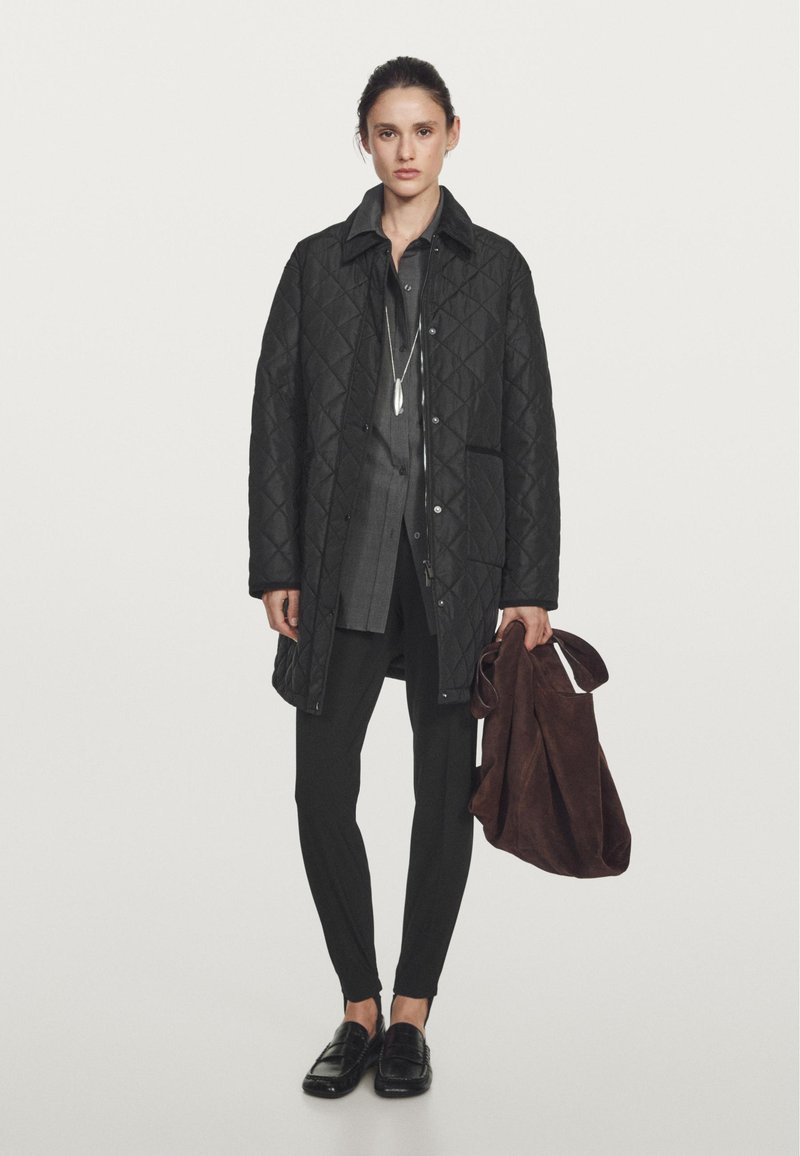 Cappotto nero trapuntato con colletto strutturato, indossato sopra una camicia grigia a maniche lunghe. Pantaloni slim neri e mocassini lucidi. Borsa a tracolla marrone tenuta in mano.