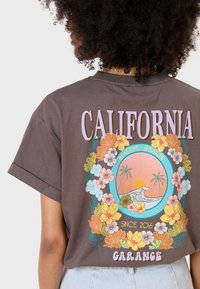 Camiseta de algodón marrón con gráficos florales coloridos, "CALIFORNIA," "MALIBU BEACH," y un diseño de ola. Manga corta y corte relajado.