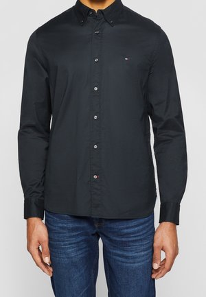 Homme portant une chemise noire à manches longues boutonnée avec col et petit logo sur la poitrine, associée à un jean bleu.