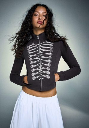 Ung kvinde med langt krøllet hår iført en sort lynlåstrøje med hvidt skeletdesign og løse hvide bukser, poserer selvsikkert.