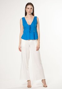 Blauwe linnen peplumtop met diepe V-hals en strikdetail, gecombineerd met witte wijde broekspijpen. Kenmerkt goudaccenten op de top.
