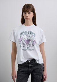 Wit katoenen T-shirt met een grafische print in paars en zwart. Korte mouwen en een ruime pasvorm. Gecombineerd met donkergrijze jeans.