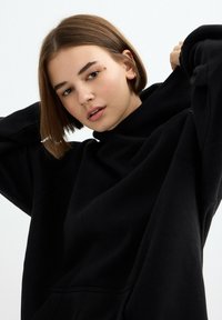 Sudadera negra de gran tamaño con cuello alto y mangas largas, fabricada con tela suave, con un bolsillo frontal y detalles mínimos.