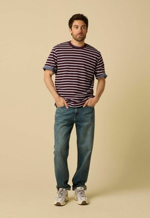Homme debout les mains dans les poches, portant un t-shirt rayé bordeaux et gris, un jean bleu et des baskets blanches sur fond beige.