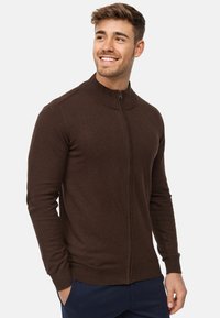 Brauner Reißverschluss-Pullover mit geripptem Kragen und Bündchen, mit tailliertem Schnitt und glatter Textur, getragen über dunkelblauen Hosen.