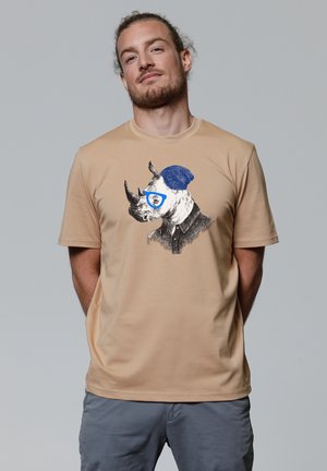 Beige katoenen t-shirt met een zwart-wit illustratie van een neushoorn die blauwe brillen en een blauwe beanie draagt. Korte mouwen, ronde hals.