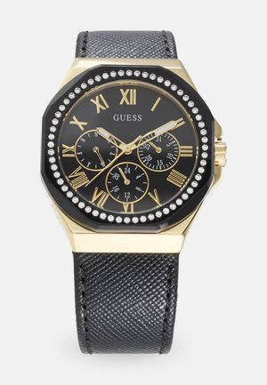 Montre octogonale Guess avec un cadran noir, des chiffres romains dorés, trois sous-cadrans et un bracelet en cuir texturé noir. Accentuée de strass.
