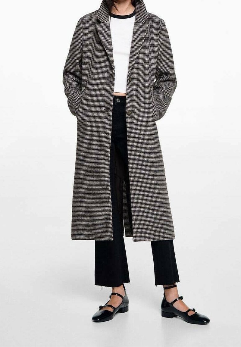 Personne portant un long manteau à motif pied-de-poule par-dessus un top court blanc et un pantalon court noir, avec des chaussures Mary Jane noires.
