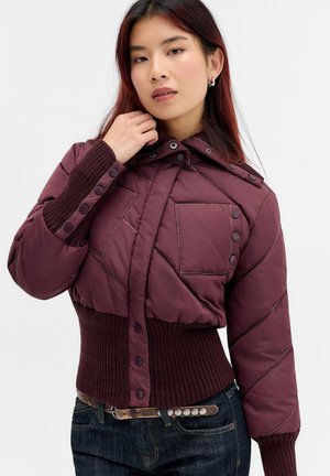 Ung kvinde iført en bordeaux quiltet jakke med knapdetaljer og ribstrikkede manchetter, kombineret med mørke jeans og et bælte med metalnitter.