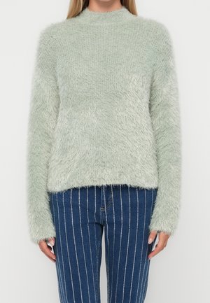 Femme portant un pull en maille pelucheux vert clair et un jean bleu taille haute avec des rayures blanches verticales, debout devant un fond blanc.