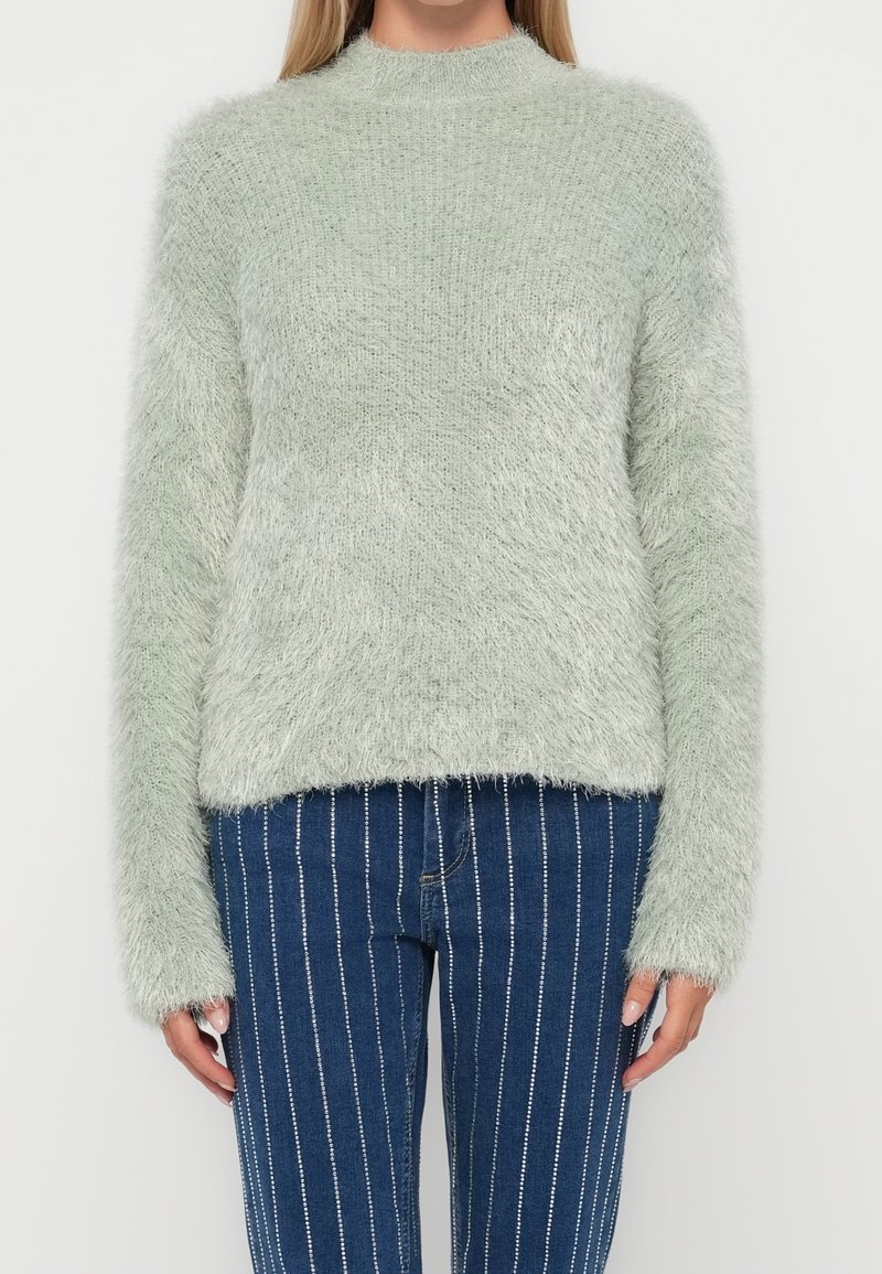 Femme portant un pull en maille pelucheux vert clair et un jean bleu taille haute avec des rayures blanches verticales, debout devant un fond blanc.