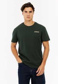 Man in een donkergroene t-shirt met "PROTEST" logo en een blauwe spijkerbroek, staande met één hand in zijn zak tegen een witte achtergrond.