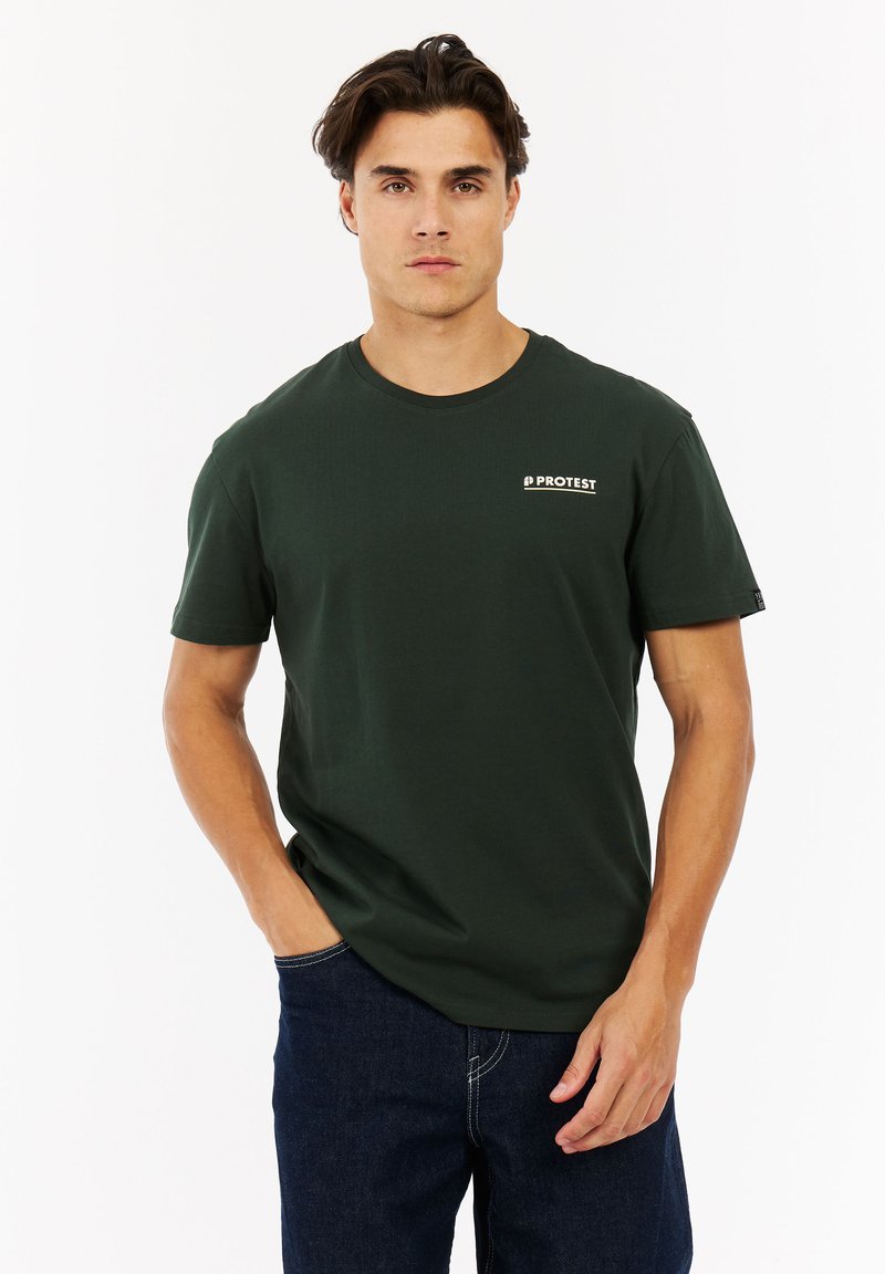 Man in een donkergroene t-shirt met "PROTEST" logo en een blauwe spijkerbroek, staande met één hand in zijn zak tegen een witte achtergrond.