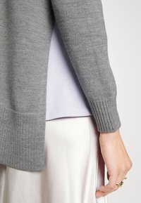 Grauer gestrickter Pullover mit gerippten Bündchen, über einem hellvioletten Top getragen, kombiniert mit einem seidigen, hellen Rock.