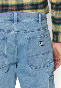 Ljusblå denim cargobyxor med två stora bakfickor, synlig sömnad och en svart logotypmärke på den högra fickan.