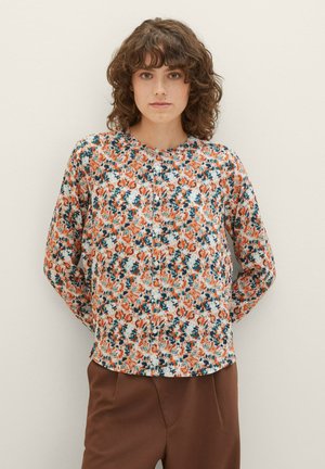 Blouse florale avec une base blanche ornée de motifs orange, bleu et vert, à manches longues et col rond avec fermetures à boutons.
