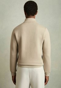Beige ribbet striktrøje med høj krave, lange ærmer og tekstureret stof, parret med lyse bukser, set fra ryggen.