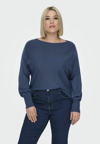 Modra, sproščena pulover z širokim ovratnikom in rebrastimi manšetami, pravilno kombiniran s temno modrimi kavbojkami. Tkanina se zdi mehka in teksturirana.
