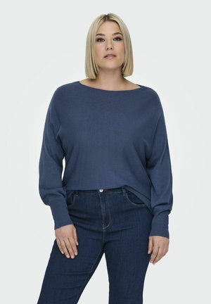 Maglione blu, dal taglio ampio, con scollatura ampia e polsini a coste, abbinato a jeans in denim scuro. Il tessuto sembra morbido e dalla texture interessante.