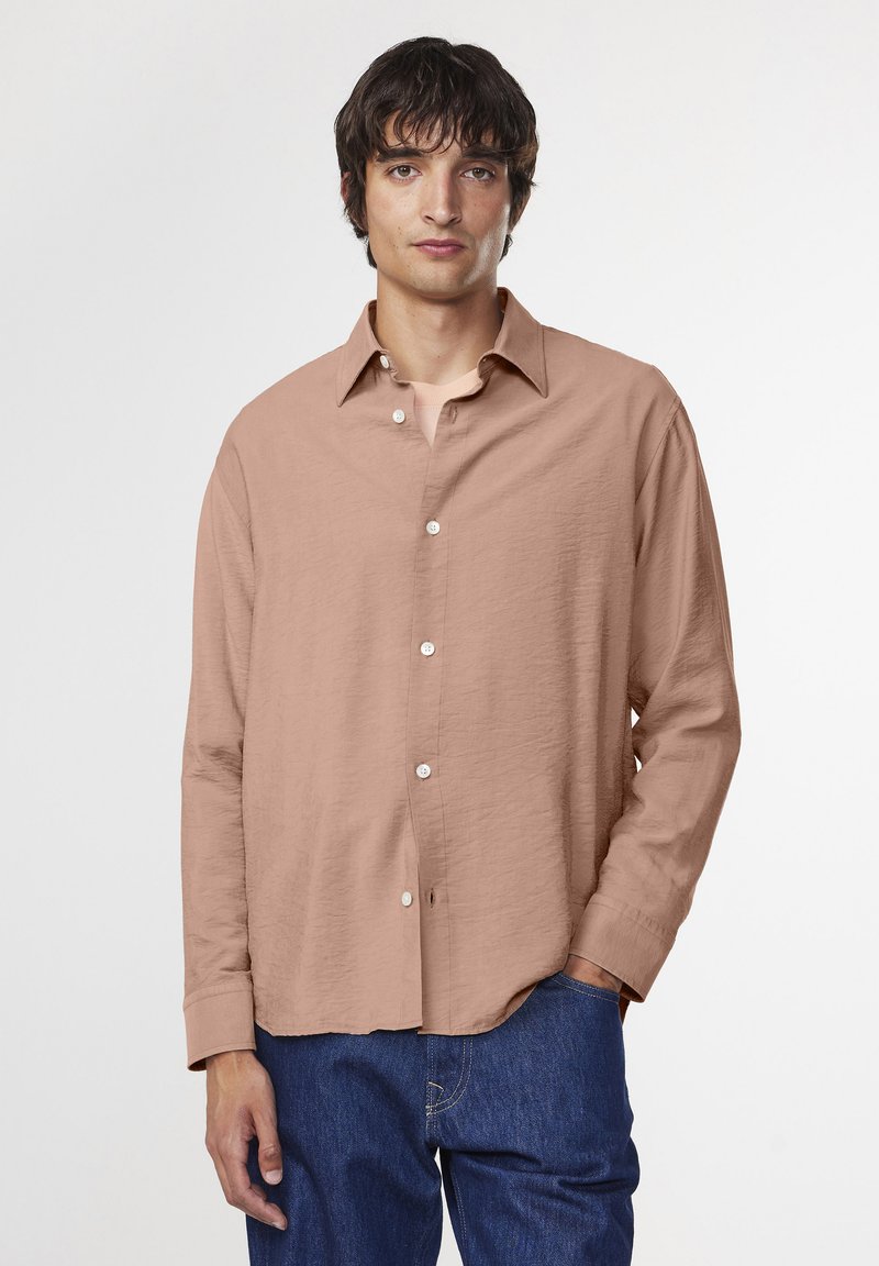 Camicia a maniche lunghe color marrone chiaro con una sottile texture increspata, chiusura con bottoni e colletto classico. Abbinata a pantaloni in denim blu.