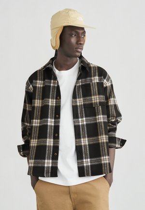 Νεαρός άνδρας με μπεζ χειμερινό καπέλο, πουκάμισο καρό σε μαύρο και μπεζ πάνω από λευκό μπλουζάκι, και μπεζ παντελόνι, κοιτάζει προς τα πλάγια με τα χέρια στις τσέπες.