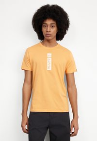 Kortærmet orange t-shirt lavet af bomuld, med et hvidt vertikalt "ALPHA" grafisk design centreret på brystet og et mærke på venstre ærme.