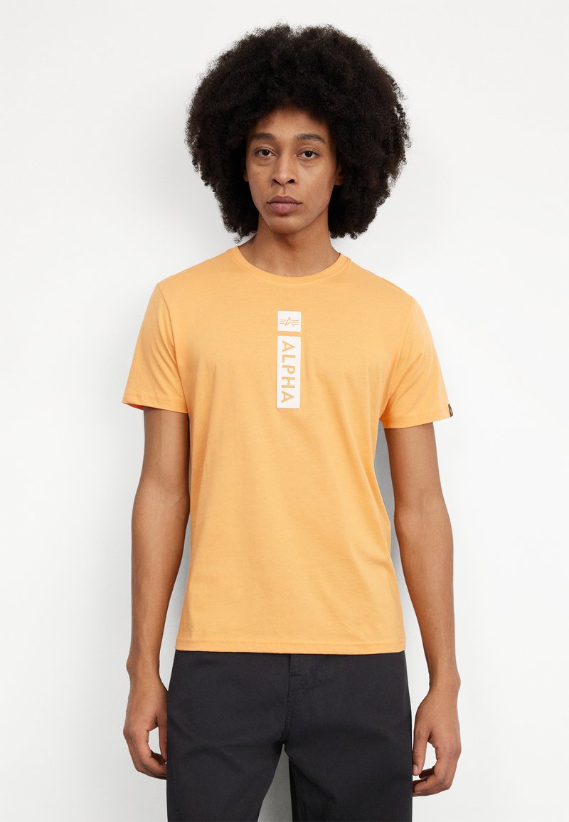 Camiseta de manga corta color naranja hecha de algodón, con un gráfico blanco vertical de "ALPHA" centrado en el pecho y una etiqueta en la manga izquierda.
