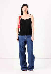 Top negro con pliegues sin mangas combinado con jeans de mezclilla azul de tiro alto y bolso rojo de hombro. La modelo se encuentra de pie contra un fondo liso.