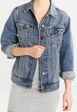 Person iført en blå denimjakke med opkrullede ærmer over en lys grå kjole, der holder jakken lukket med den ene hånd.