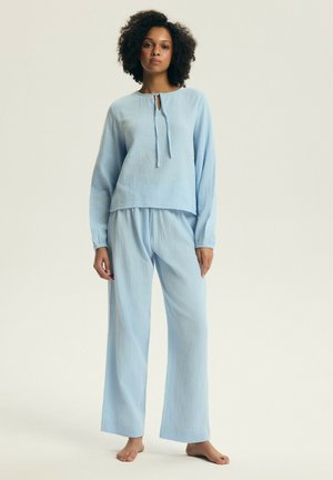 Kobieta stojąca boso, ubrana w luźną, jasnoniebieską, teksturowaną bluzkę z długim rękawem i spodnie o szerokich nogawkach na jednolitym tle.