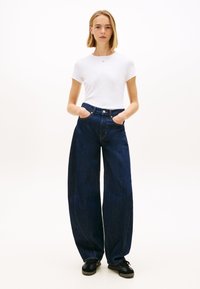 Biała koszulka z krótkim rękawem w połączeniu z szerokimi spodniami dżinsowymi w ciemnym odcieniu. Spodnie mają zapięcie na guziki i boczne kieszenie. Na nogach czarne buty.